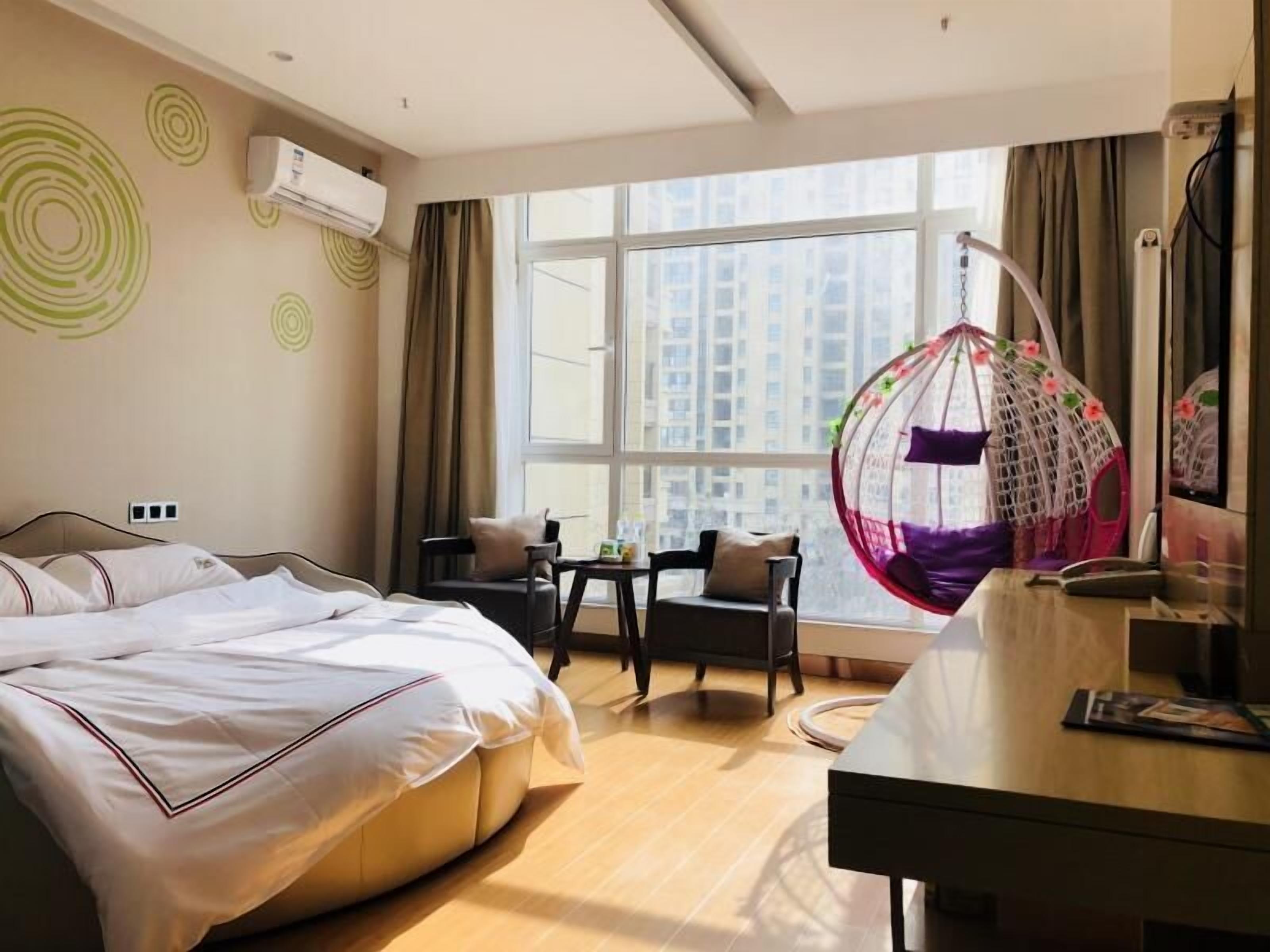 A hotel room with a bed, a desk, a chair, a hanging chair, and a large window with a city view.