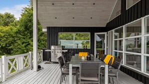 En udendørs terrasse med grill, siddepladser og store vinduer.