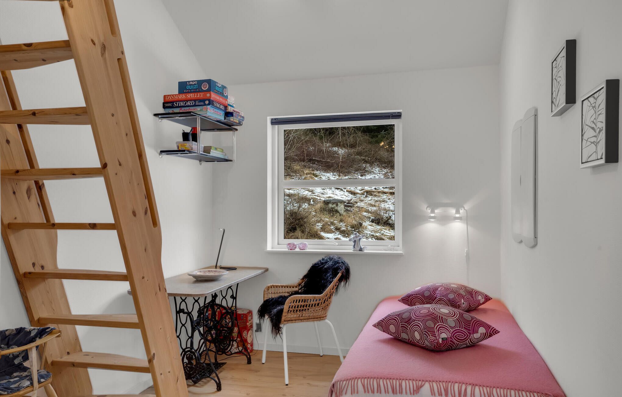 Ett litet, ljust rum med en trästege, ett skrivbord med böcker, en stol, en säng med mönstrade kuddar och ett fönster med utsikt över ett snötäckt landskap.