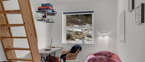 Ett litet, ljust rum med en trästege, ett skrivbord med böcker, en stol, en säng med mönstrade kuddar och ett fönster med utsikt över ett snötäckt landskap.