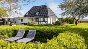 Une maison avec un toit en pente, un jardin agrémenté d’une haie et deux fauteuils de jardin.