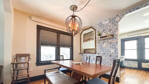 A dining area with a wooden table, benches, and chairs. There is a chandelier above the table, and a window with blinds.