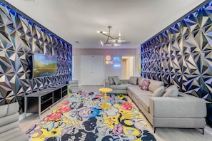A modern living room with a geometric patterned wall, a colorful cartoon-themed rug, and a grey sofa.