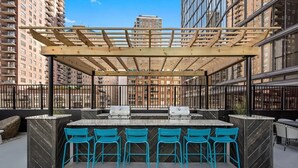 Un espace bar extérieur doté d’une pergola en bois, d’un comptoir en marbre et de chaises bleues.