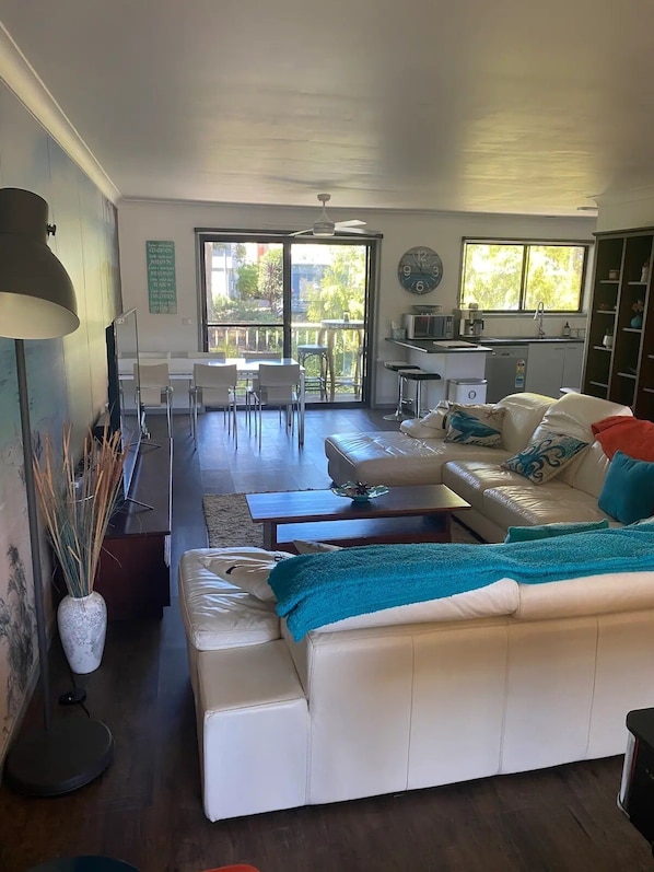 A spacious living area with a white sofa, a dining table, and a kitchen in the background. - Denhams Beach House (with tennis court/pool/wifi!) (Denhams Beach)