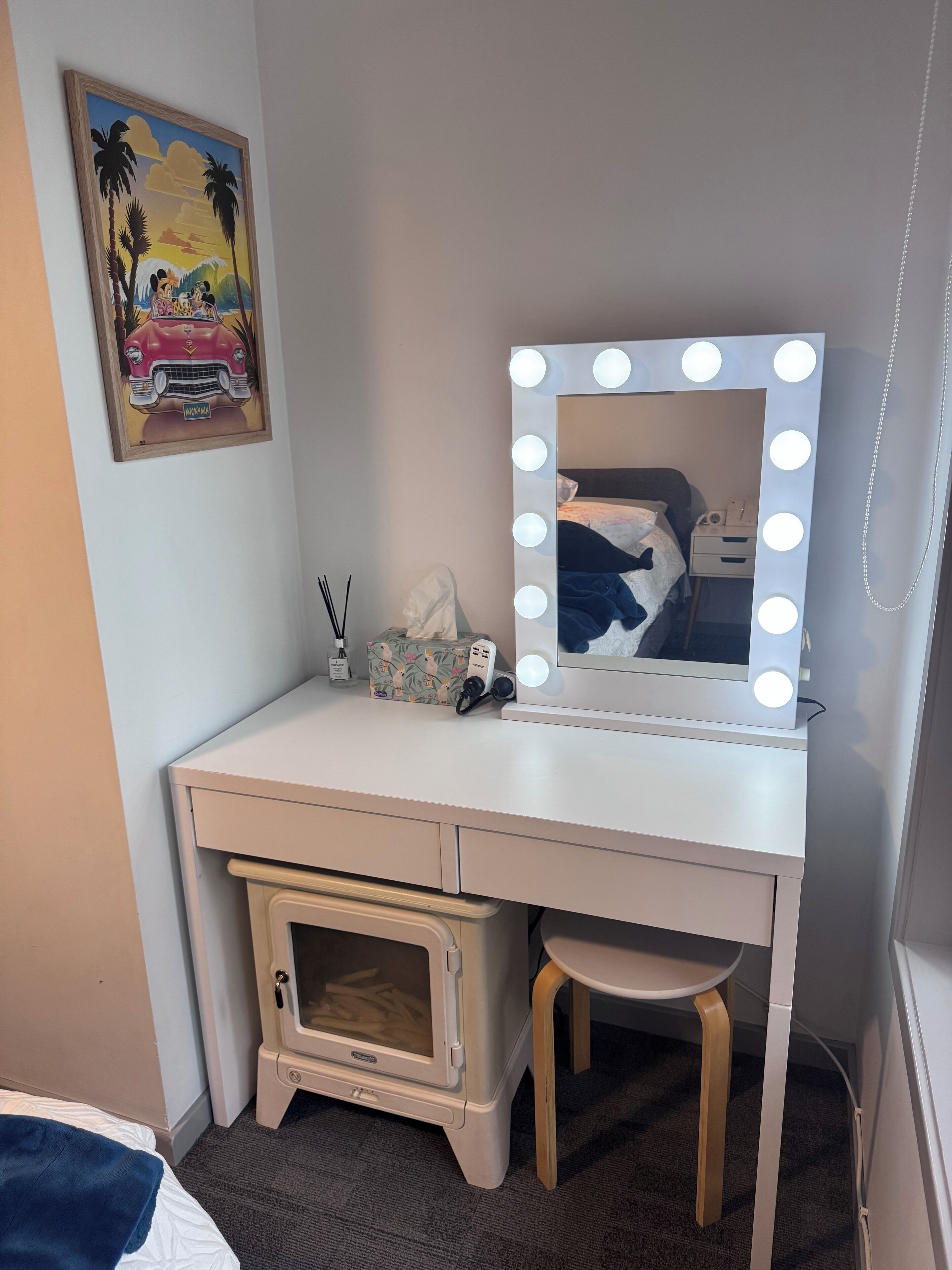A white vanity with a mirror surrounded by lights, a small stool, a tissue box, and a bottle.