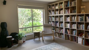 A room with a large window, a bookshelf filled with books, a chair, a drum set, and a potted plant. - Doreen Gem (Doreen)