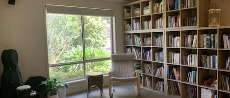 Une pièce avec une grande fenêtre, une bibliothèque remplie de livres, un fauteuil, une batterie et une plante en pot.