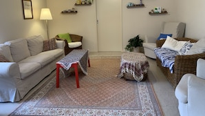 A living room with a beige sofa, a wicker chair, a patterned ottoman, a floor lamp, and a decorative rug. - Doreen Gem (Doreen)