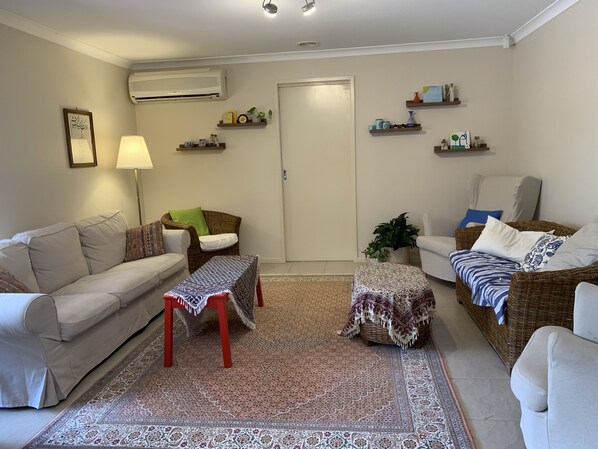 Ein Wohnzimmer mit beiger Couch, Korbsessel, gestreiftem Hocker, Stehlampe und einem dekorativen Teppich.
