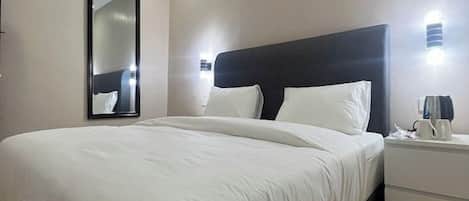 A hotel room with a large bed, a nightstand with a lamp, a mirror, and a wall-mounted light fixture.