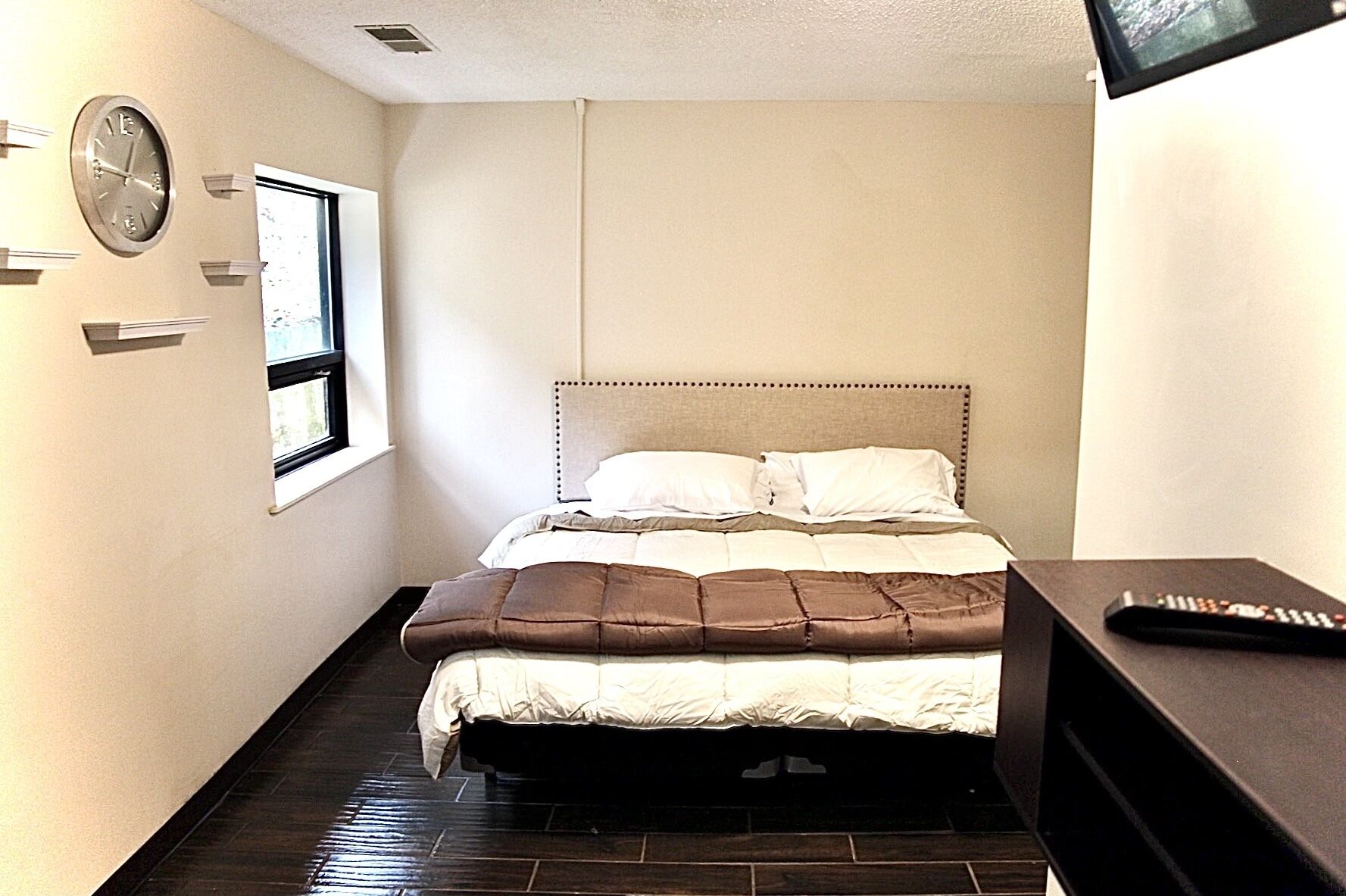 A bedroom with a bed, a clock, a window, and a remote control on a table.