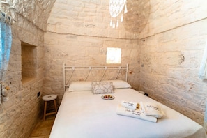 Dormitorio con paredes de piedra, una cama, un taburete y una ventana.