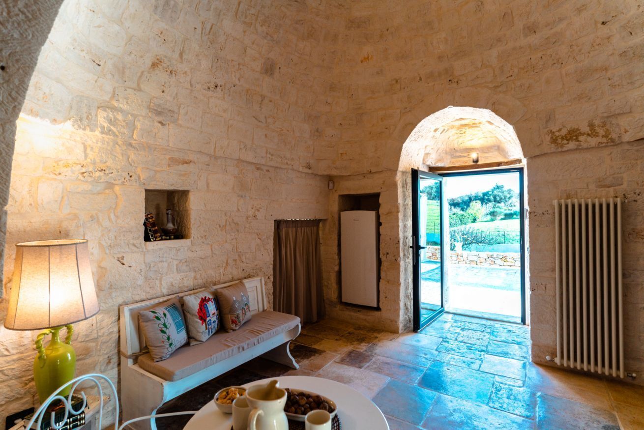 A stone-walled room with a sofa, a table with a pitcher and bowls, and a view through an open door to an outdoor area.