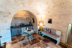 A stone-walled room with a kitchen area, a round table with chairs, and a bench with cushions.
