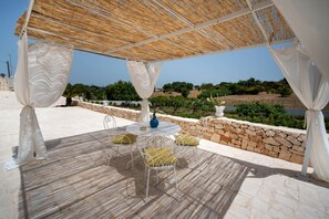 An outdoor patio with a stone floor, white metal chairs with yellow cushions, a round table with a blue vase, and a view of the landscape. - WePuglia - Trullo Meraviglioso (Martina Franca)