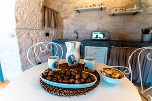 A rustic kitchen with a stone wall, a white metal chair, a table with a blue plate of nuts, two ceramic cups with rooster designs, and a vintage toaster.