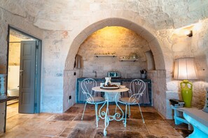Una habitación rústica con paredes de piedra, una mesa redonda y dos sillas, una pequeña cocina con un radio, y una puerta que da a otra habitación.
