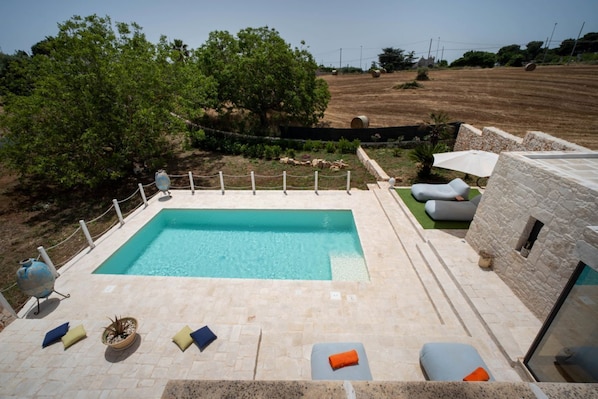 Una piscina con solárium de piedra, rodeada de sillones de descanso y un muro de piedra.