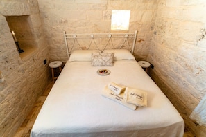 Une pièce étroite aux murs de pierre, avec un lit simple, une petite fenêtre et une bougie posée sur la table de chevet.