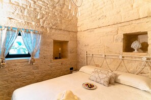 Une chambre aux murs de pierre, avec un lit, une fenêtre avec des rideaux et une petite étagère sur laquelle est posé un objet décoratif.