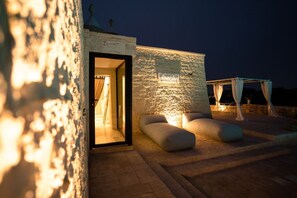 Een gebouw van steen met een verlichte ingang, terrasplekken en een bord.