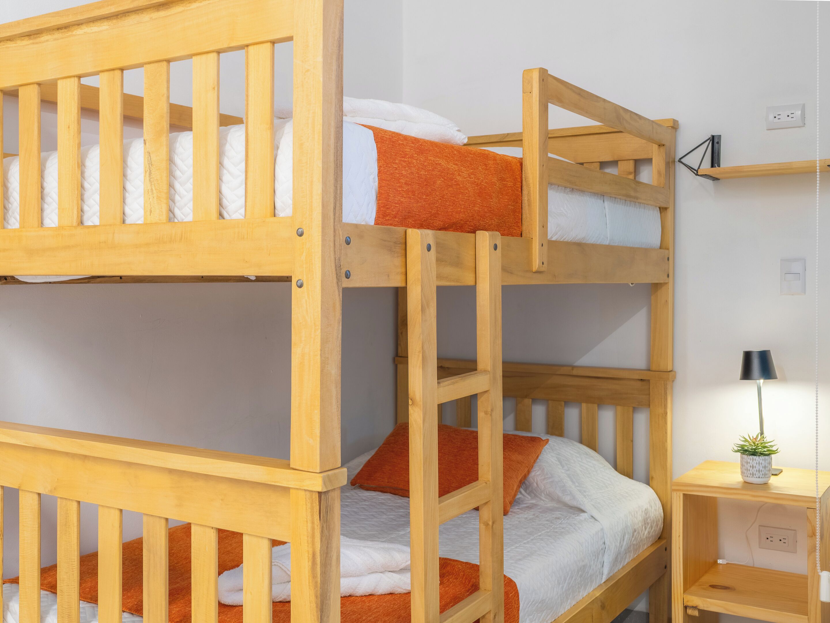 Un lit superposé avec une structure en bois, une literie blanche et des oreillers orange. On y trouve une petite table de chevet en bois, sur laquelle se trouve une lampe et un pot de fleurs.