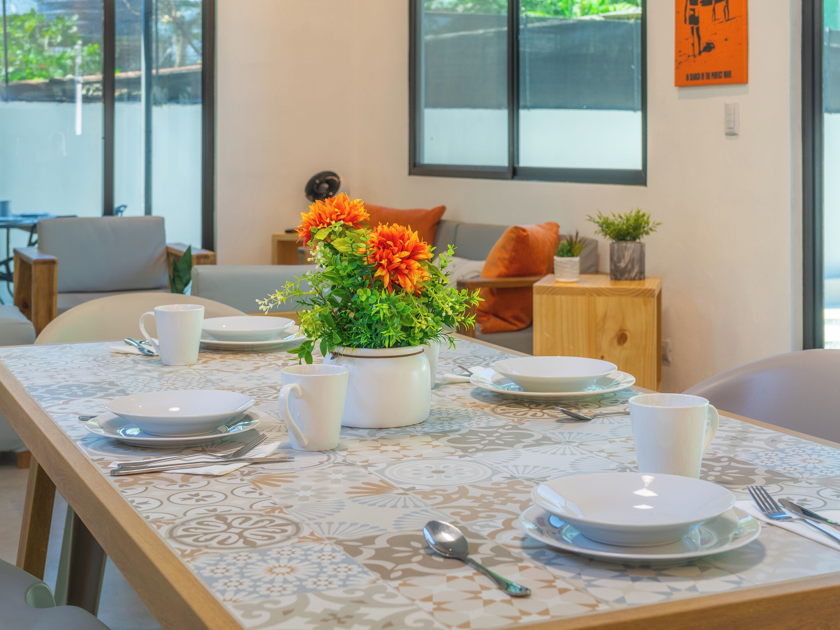 A dining area with a table set for four, a vase with orange flowers, and a view of the outdoors through large windows.