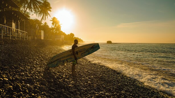 A person holding a surfboard on a pebbly beach with palm trees and a sunset in the background. - El Salvador Surf Houses (La Libertad)