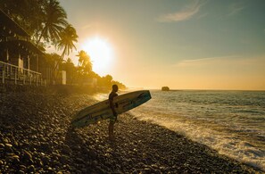 A person holding a surfboard on a pebbly beach with palm trees and a sunset in the background. - El Salvador Surf Houses (La Libertad)