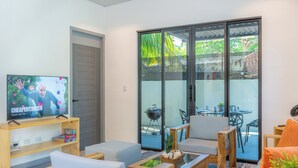 A modern living room with a TV, wooden furniture, and a sliding glass door leading to an outdoor patio. - El Salvador Surf Houses (La Libertad)