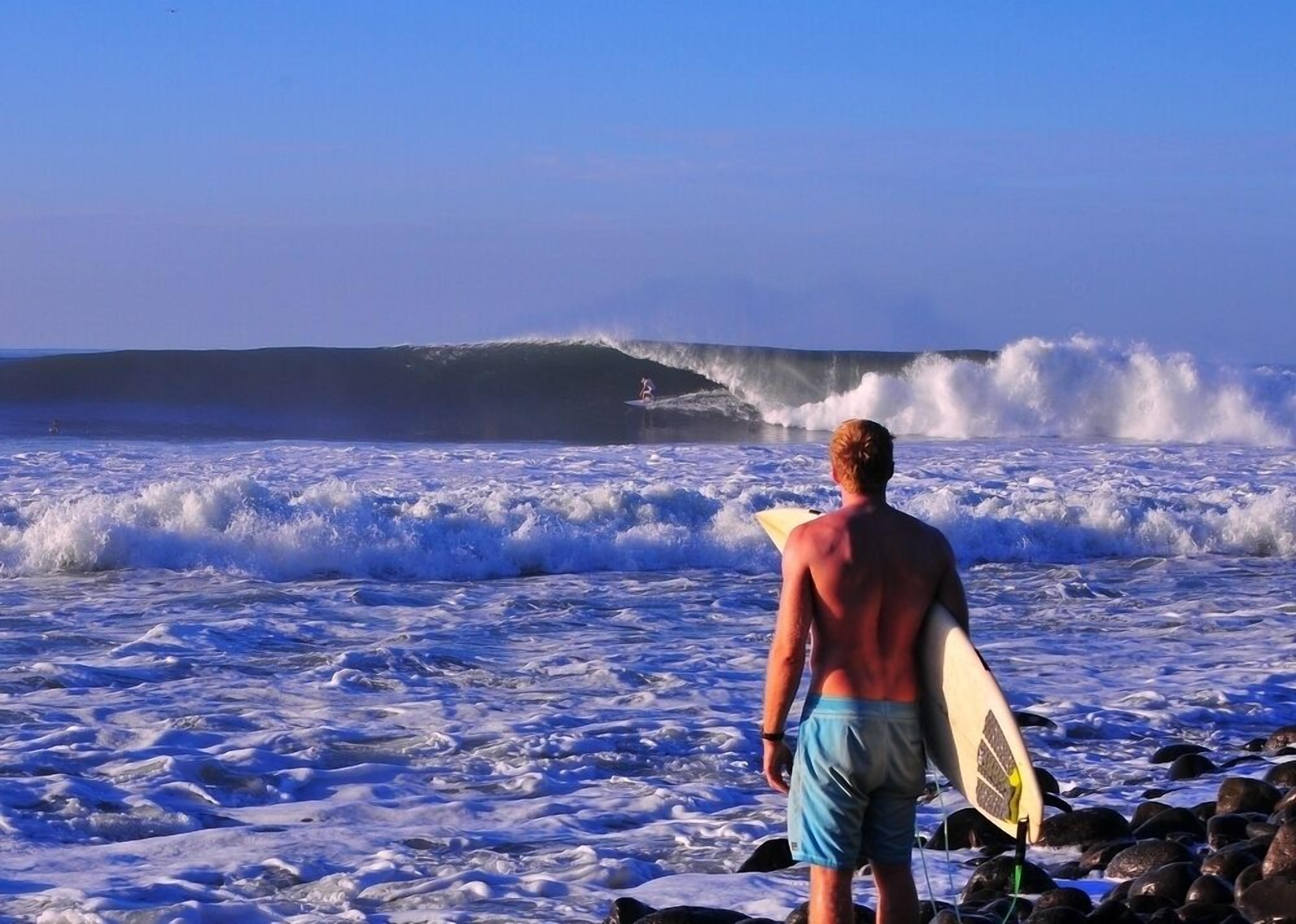 Un hombre con una tabla de surf en una playa rocosa, mirando al océano con olas rompiendo.