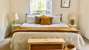 Un dormitorio con una cama grande, dos mesitas de noche con lámparas, un banco de madera y una ventana con vistas a árboles.