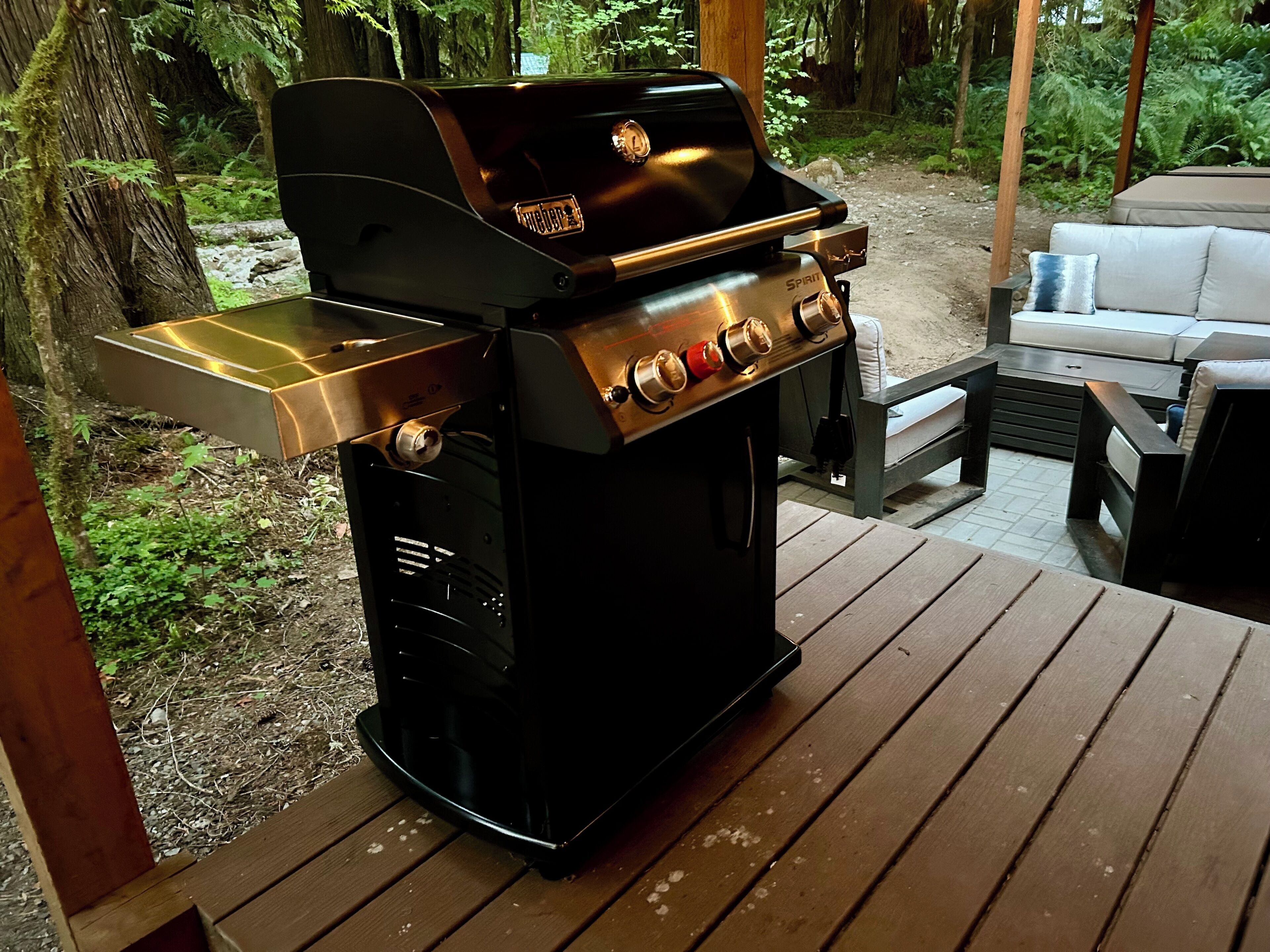 Una parrilla de barbacoa en una terraza de madera, con un área de estar exterior acolchada al fondo.