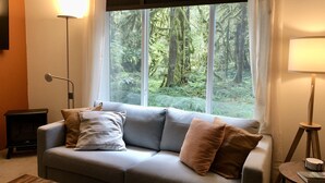 Una habitación con un sofá gris, dos cojines, una lámpara de pie, una mesita con lámpara y una ventana con vistas a un bosque.