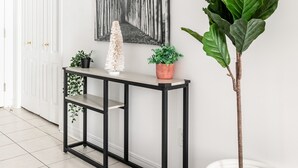 Een hal met een witte deur, een zwarte console tafel met planten, een grote potplant en een zwart-wit abstract schilderij aan de muur.