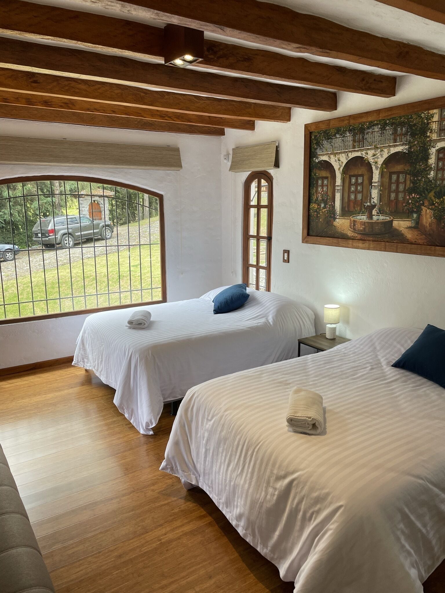 Una habitación con dos camas, un cuadro, una ventana que da a la vista de un coche y vigas de madera en el techo.
