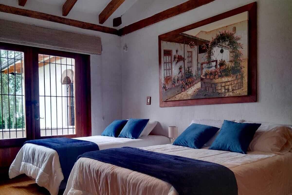 Ein Zimmer mit zwei Betten, Holzbalken an der Decke, einem großen Fenster mit Gitter und einem Gemälde an der Wand.