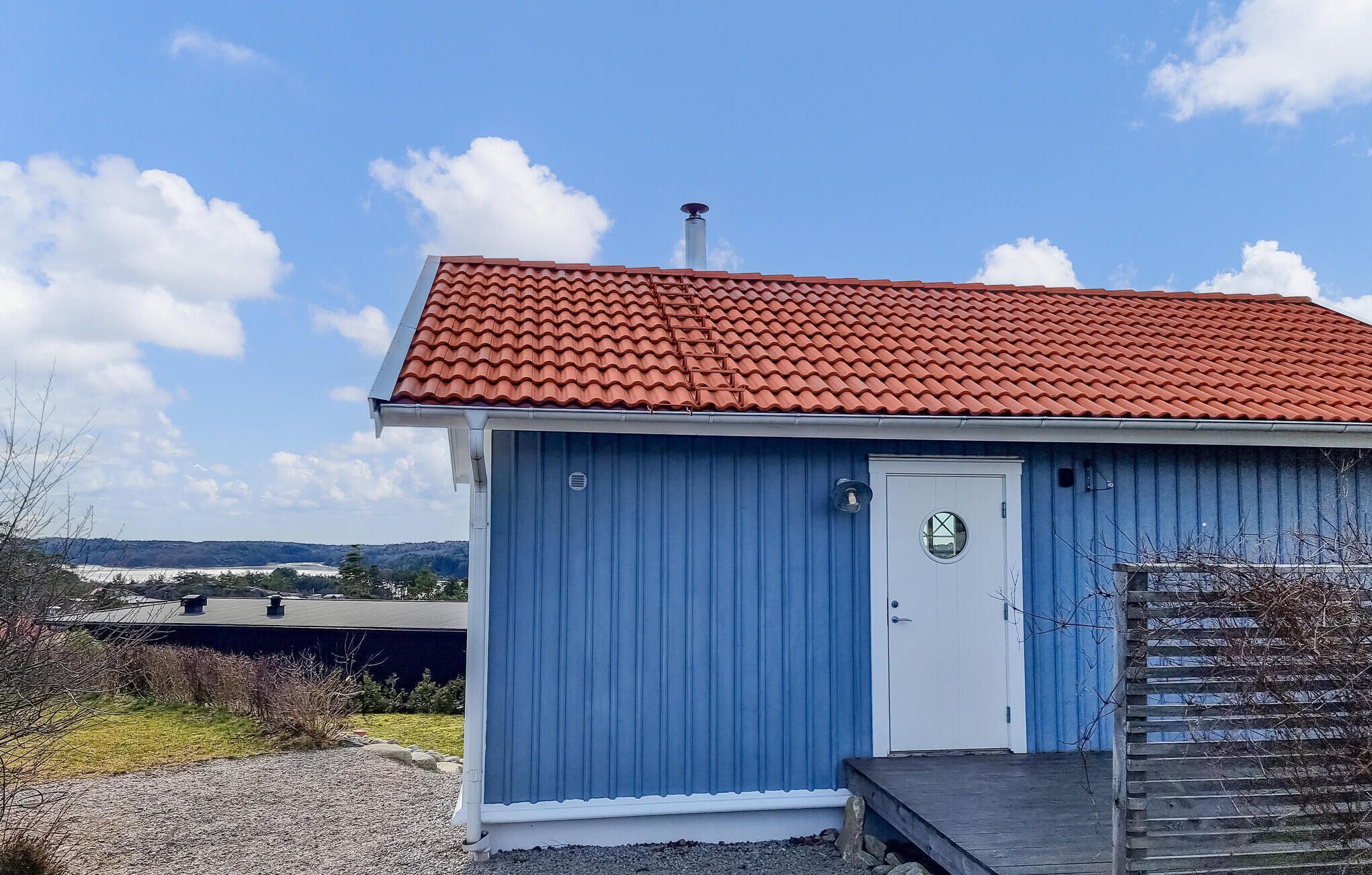 A small, single-story building with a red-tiled roof and a white door, surrounded by a gravel driveway and sparse vegetation.