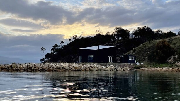 A modern house with a flat roof, situated on a rocky shoreline with a body of water in the foreground.