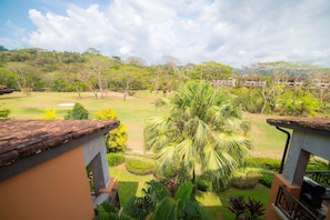 A view of a golf course with a large palm tree, a building with a tiled roof, and a hill with trees and a house in the background.