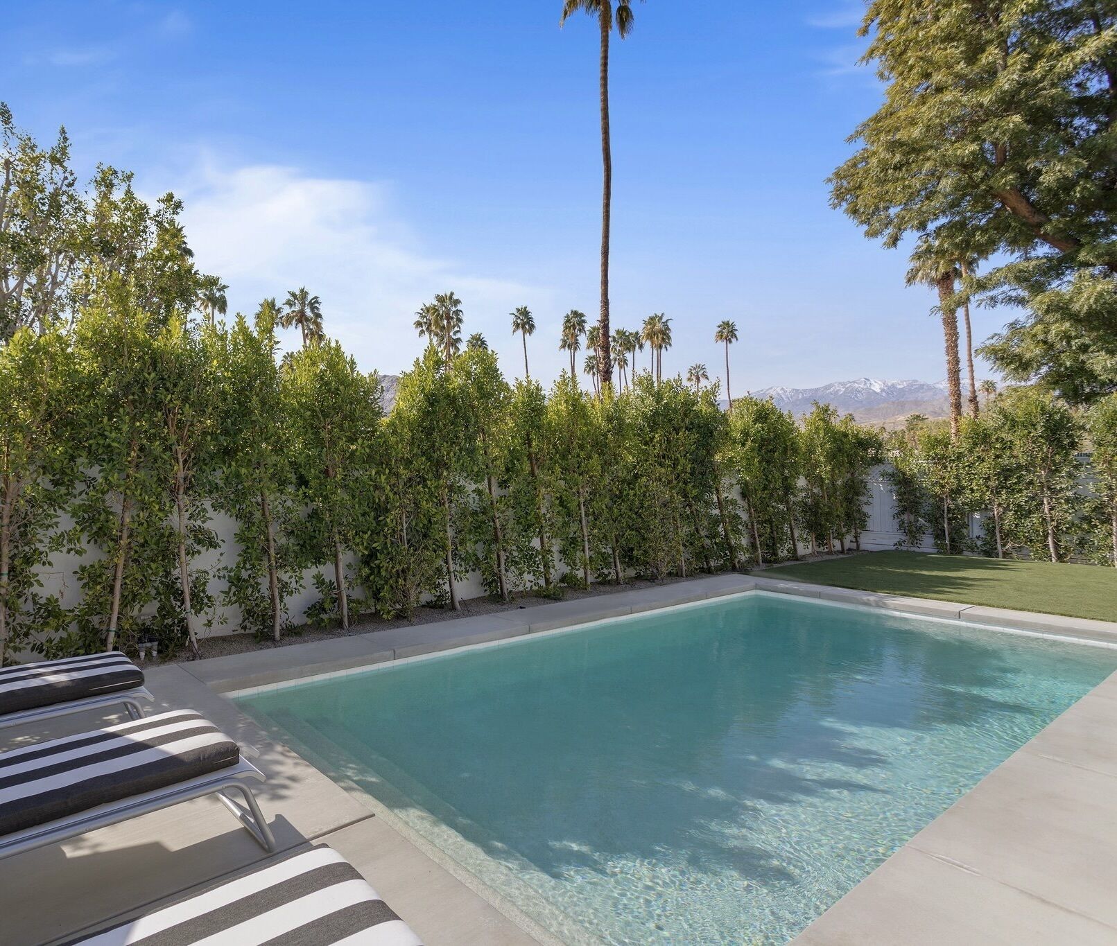 A rectangular swimming pool with clear blue water, surrounded by a low wall with various trees and shrubs. There are lounge chairs with striped cushions along the poolside.