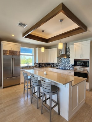 A modern kitchen with a large island, bar stools, stainless steel appliances, and a view to the outdoors.