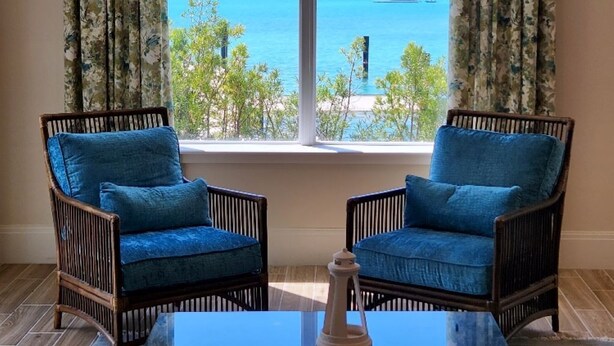 A room with two wicker chairs with blue cushions, a glass coffee table, and a view of a body of water through a large window with floral curtains. - Two Bedroom Luxury Condo located in Beautiful February Point (Exuma)