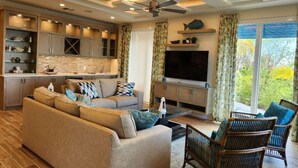 A modern living room with a sofa, chairs, a TV, and a kitchen area with cabinets and a countertop. - Two Bedroom Luxury Condo located in Beautiful February Point (Exuma)