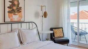 Une chambre à coucher avec un lit, une petite table, une chaise et une fenêtre donnant sur des immeubles.