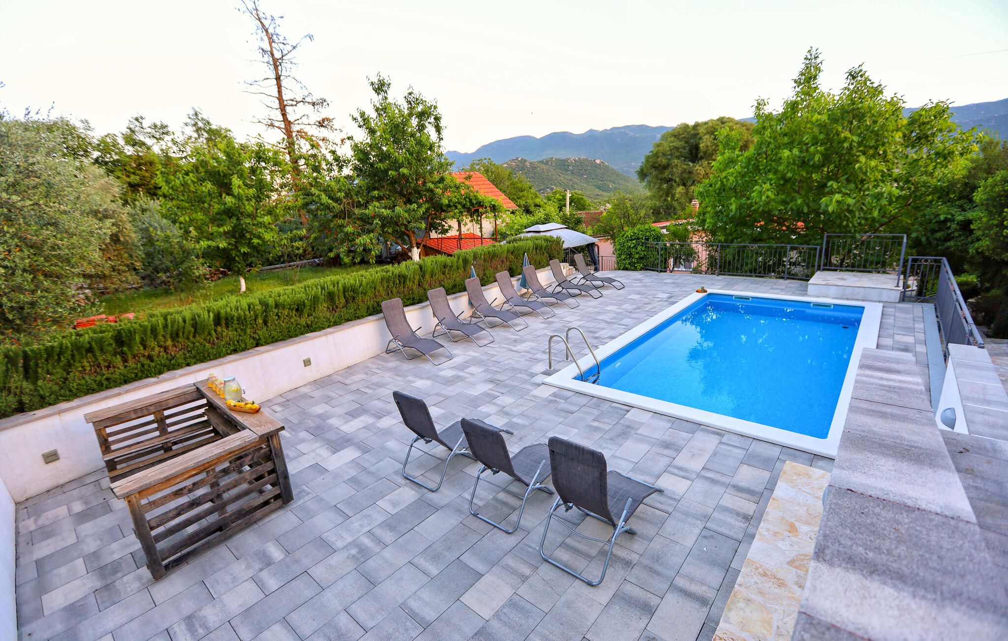 Une piscine entourée de chaises longues et d’un banc en bois, elle-même bordée d’un jardin aménagé et avec des montagnes en toile de fond.