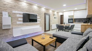 Ein modernes Wohnzimmer mit einem Flachbildfernseher, grauen Sofas, einem Holztisch und einer Küchenzeile mit weißen Schränken und einer schwarzen Arbeitsplatte.