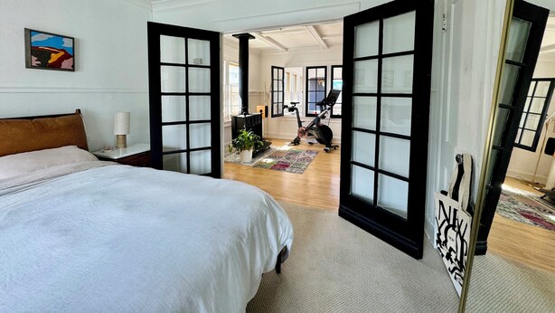 A bedroom with a bed, a mirror, a door leading to a living area with a fireplace, and exercise equipment. - Historic & sunny beachside 2br with modern comforts (Marina del Rey)