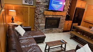 Uma sala de estar aconchegante com lareira de pedra, sofá de couro em formato de L, mesa de madeira e televisão fixada na parede.
